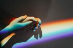 Photo of two white hands, each from a different person, holding the other gently in front of a refracted rainbow on the white wall behind them