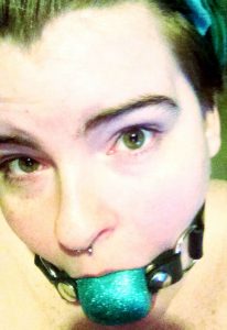Photo of Sugarcunt in a glittery turquoise silicone ball gag. They are looking up at the camera with brows raised and hazel eyes wide with anticipation.