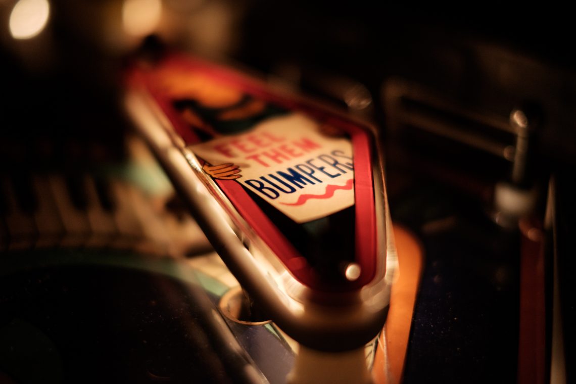 A macro photo of a bumper slingshot on a pinball machine - the bumper is chrome with a red band and a sign that reads, "Feel them bumpers," with "bumpers" underlined. The sign appears to be held by a character with a beard, but the rest of the character is out of the light and focus.