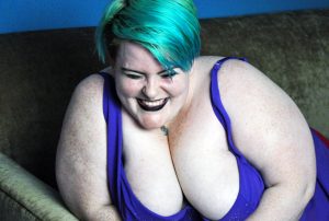 Photo of Sugarcunt, a white, fat, assigned female at birth person with teal hair, black lipstick, and facial piercings. They are in the middle of laughing, and have a huge smile on their face.
