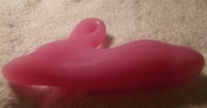 A pink dildo sits on a white background.  The dildo is relatively phallic, with a bulbous bump near the head of the toy.  A protruding, handle-like piece is positioned at the bottom of the toy, complete with a hole that would be ideal as a fingerhold