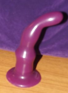 The Tantus protouch sits on a wooden desk in front of a purple microfiber bag. The protouch is wine-colored, with a stout base that flares out, then tapers into the shape of a curved finger.