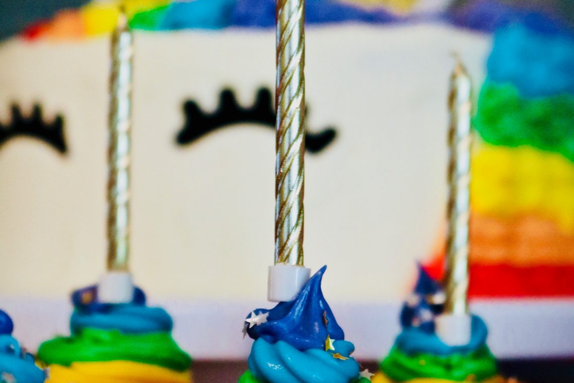Photo by James Lee on Unsplash of a large rainbow unicorn cake sitting, out of focus, behind several small rainbow-icing-bedecked cupcakes with unlit golden candles.