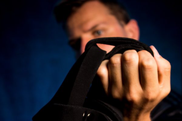 Photo of an AMAB person holding up a black harness in front of a blue background. Their face is largely obscured by the fist and harness, and their tightly-balled fist on the harness is the focus of the photo.