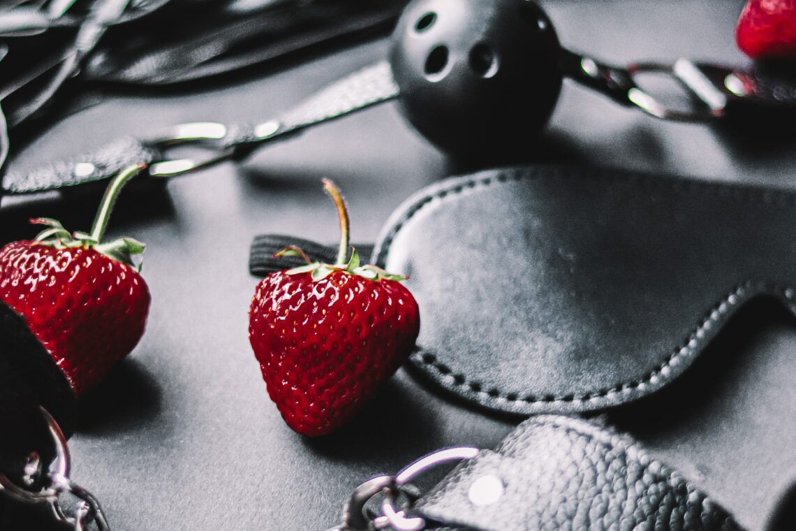 Black and white photo of bondage cuffs, a flogger, a blindfold, a ball gag, and red strawberries placed around them.