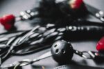 Black and white photo of bondage cuffs, a flogger, a blindfold, a ball gag, and red strawberries placed around them.
