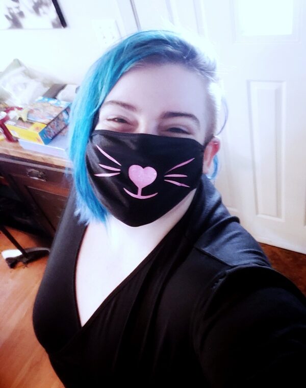 Photo of Sugar smiling brightly with blue, freshly-side-shaved hair in a black dress. They wear a black face mask with a cute pink heart nose, mouth, and whiskers on it in the likeness of a cat's face. The mask is from pinkKittee.com