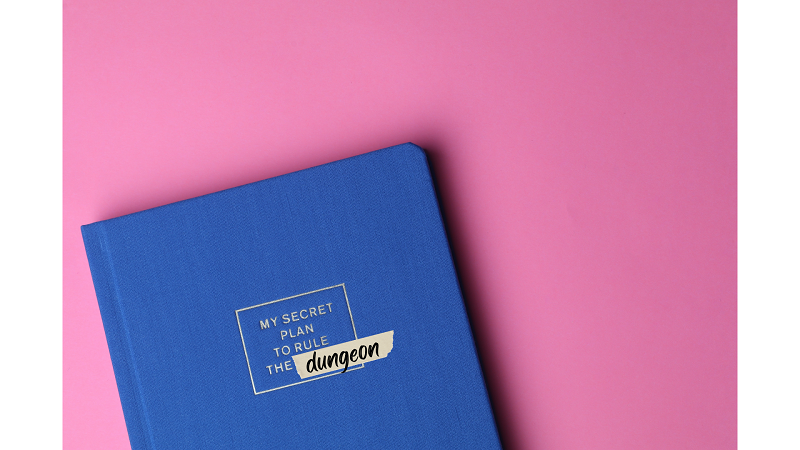 Photo of a blue planner cover with embossed gold lettering reading: my secret plan to take over the dungeon," "dungeon" is written on a piece of tape covering the original embossed word beneath