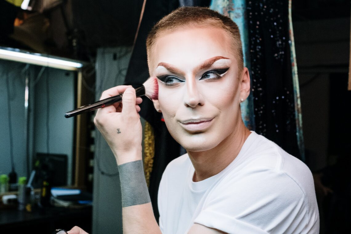 Photo of a white person applying pale white base foundation and a dark cheekbone contour - they appear to be getting ready backstage somewhere.