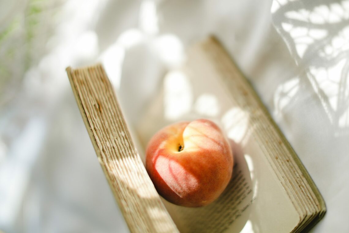 Photo of an old book lying in the sunlight, its pages held open by a whole peach sitting in the middle of the book
