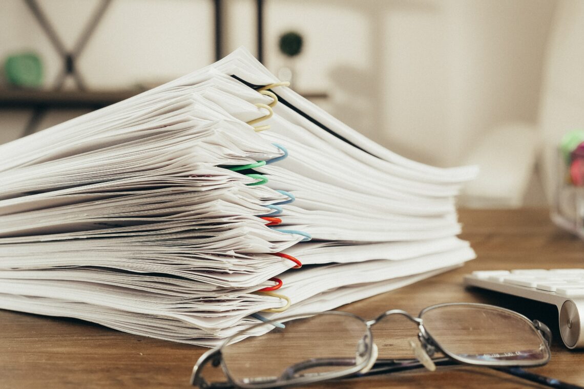 Photo of a stack of several binder-clipped paper bundles stacked beside a pair of glasses on a table