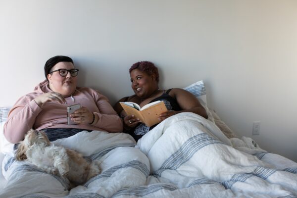 Photo of two fat people in bed - a white person with glasses and short, dark hair, and a Black person with close-cut brown hair. They are covered by a blanket, lounging with a white dog, talking while the Black person reads a book and the white person uses their phone.